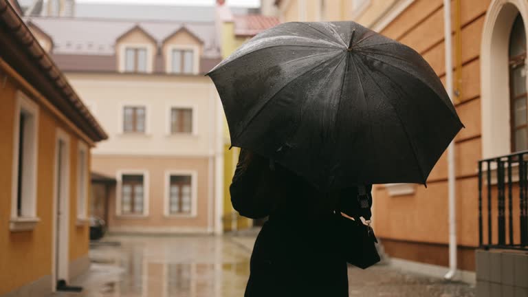 Rear view of young cute smiling attractive woman girl in coat walking alone on city street in rain, holding black umbrella and cup of coffee, turning to camera and smiling. High quality 4k footage