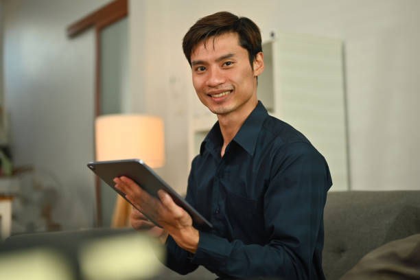 istock Smiling asian man using digital tablet, reading ebook app online or surfing internet on couch at home. 1466932910