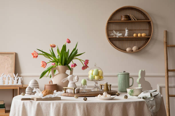 istock Creative composition of easter dining room interior with round table, vase with tulips, colorful eggs, pitcher, wooden trace, bread, hare sculpture and personal accessories. Home decor. Template. 1466936452
