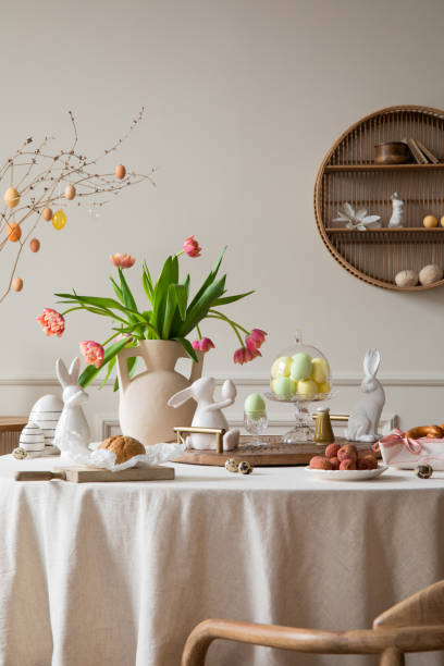 Creative composition of spring easter dining room interior with round table, vase with tulips, colorful eggs, wooden trace, bread, easter bunny, sculpture and personal accessories. Home decor Template composición creativa del interior del comedor de pascua de primavera con mesa redonda, jarrón con tulipanes, huevos de colores, rastro de madera, pan, conejito de pascua, escultura y accesorios personales. plantilla de decoración del hogar