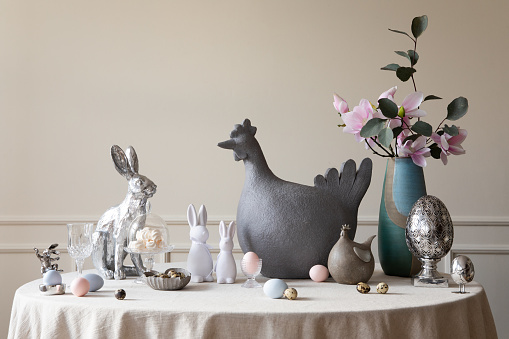Spring composition of easter dining room interior with gray hen sculpture, easter bunny, colorful eggs, vase with magnolia, beige wall with stucco and personal accessories. Home decor. Template.