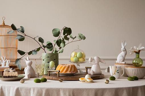 Warm and cozy composition of easter dining room interior with round table, cake, hare sculpture, colorful eggs, wooden trace, vase with leaves and personal accessories. Home decor. Template.