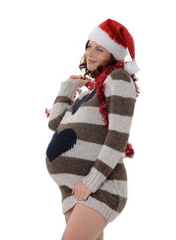 Pregnant young woman wearing santa hat and tinsel touching belly while standing over white background
