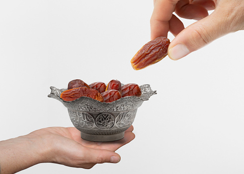 A closeup shot of a hand taking pitted dates from the vintage metal bowl on a white background