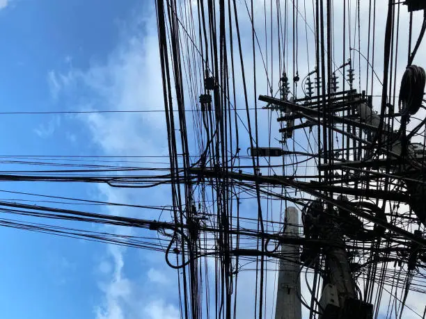 Extreme entanglement of black electric and communications cables on the pillar post at the corner of a street. Silhouettes on blue sky.