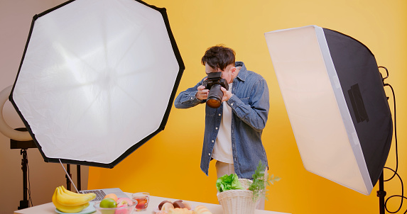 asian young male photographer use professional camera shooting food in front of yellow background at studio