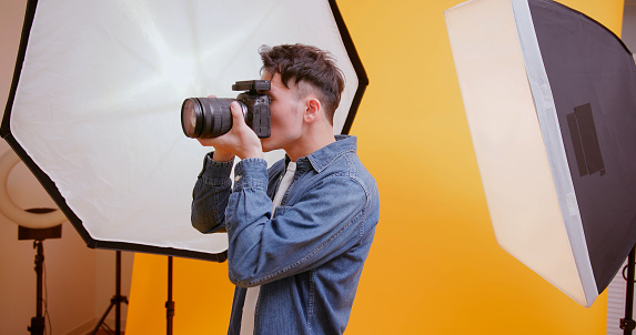 asian young male photographer holding professional camera for shooting standing in front of yellow background at studio - he feel satisfied about work