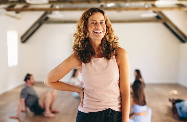 Mature woman standing in a fitness studio Mature woman smiling at the camera while standing in a fitness studio with a group of people in the background. Happy woman having an exercise session with her class in a community yoga studio. yoga instructor stock pictures, royalty-free photos & images