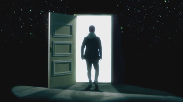Back , rear view of man walking in front of an open door with darkness behind . Footage of person walking on floor . White light inside the door. Concept of anonymous,  spirit in coma or teleportation