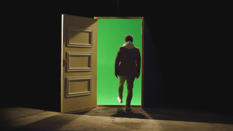 Back view of a man walking in front of an open door with darkness behind . Footage of businessman walking on floor near door with green screen . Concept of anonymous,  business dreams or teleportation