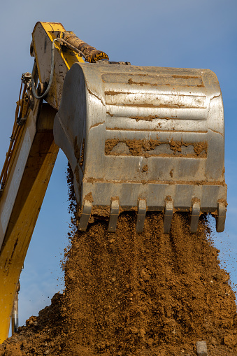 Large shining steel bucket of yellow hydraulic excavator boom unloading brown soil on construction site