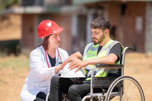 A female doctor and construction worker with disability are working together at the construction site. Construction worker with disability on wheelchair. They are talking about an accident.