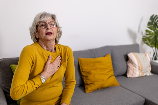 An older woman is sitting on the sofa in the living room of her apartment and holding her chest due to pain that she constantly feels.  Heart attack and chronic illness concept.