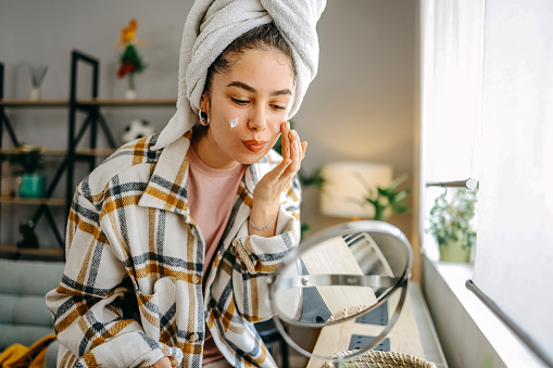 One woman, a beautiful young woman sitting in the living room applying face cream, has a towel on her head.