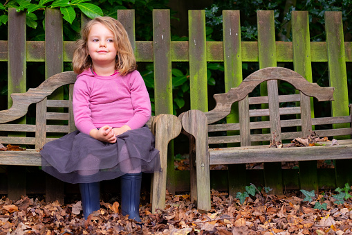 An adorable three year old girl sits on a bench in a garden.