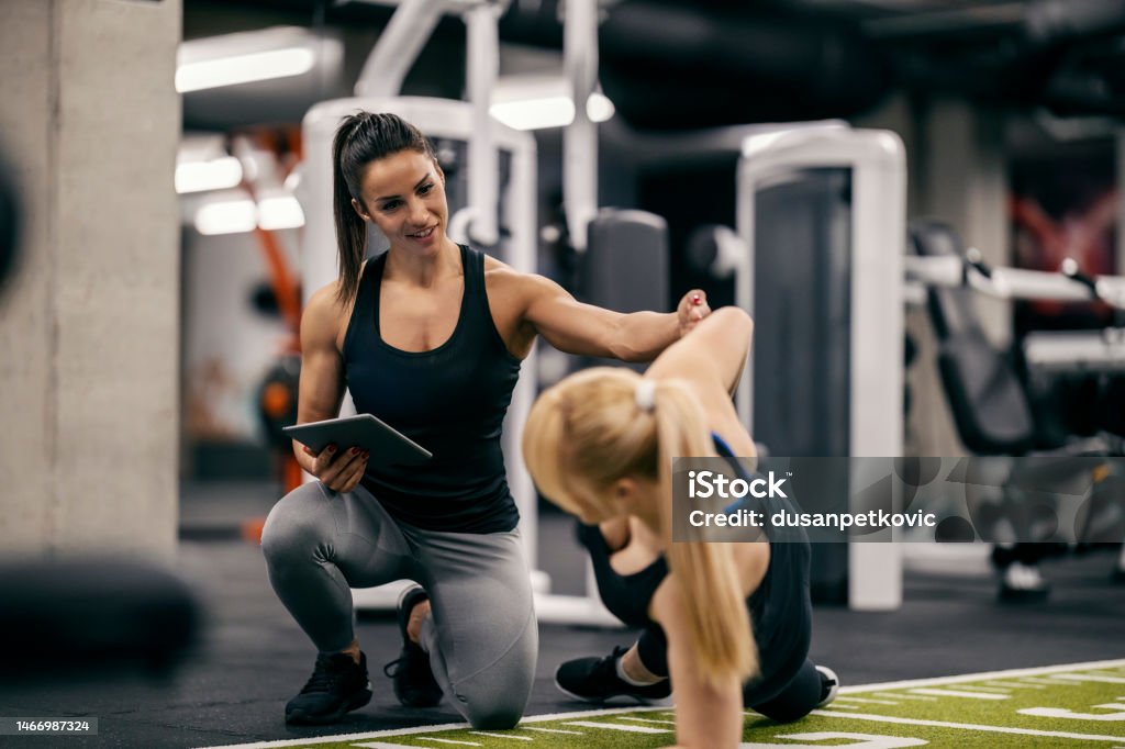 A female trainer is helping sportswoman with workouts in a gym. A female personal trainer is training sportswoman in a gym and tracking her progress on a a tablet. Coach Stock Photo