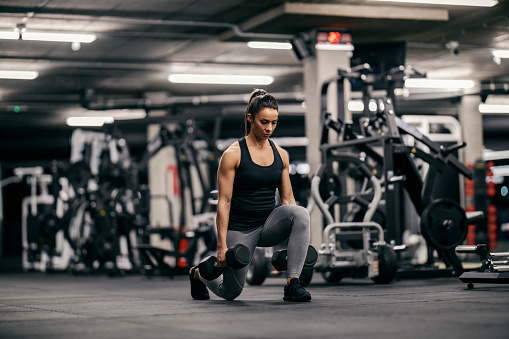 A fit female bodybuilder is lifting weights and dumbbells while doing lunges in a gym during her strength training.