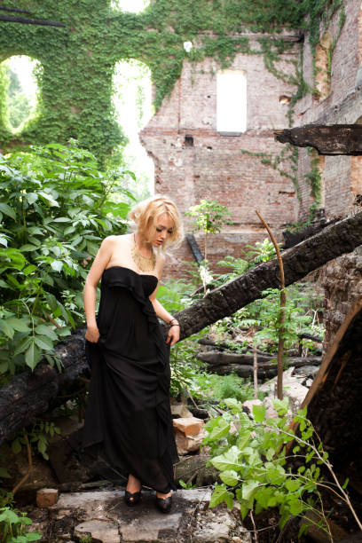 Delicate blonde girl dressed in black long dress looking down at her shoes that don't suit the harsh environment.
