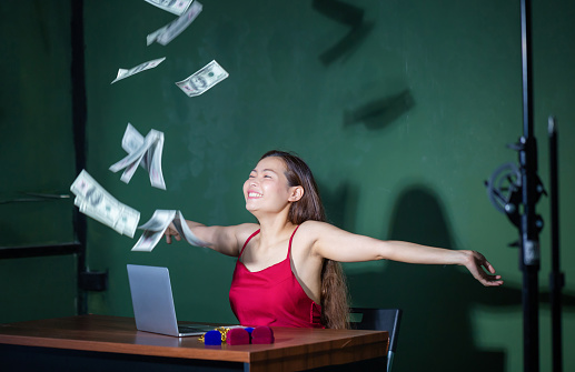Excited happy young woman sitting at table with dollars money flying overhead while raising hands