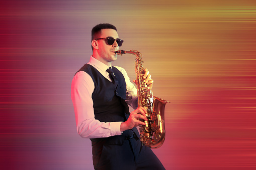 Young man in elegant outfit playing saxophone on color background
