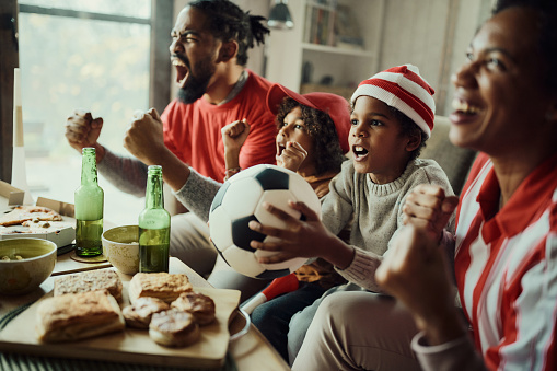 Cheerful African American family celebrating success of their favorite soccer team while watching a game on TV at home. Focus is on boy.