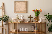 Interior design of spring living room interior with mock up poster frame, glass vase with tulips, wooden sideboard, hare sculpture, bowl, ladder, and personal accessories. Home decor. Template