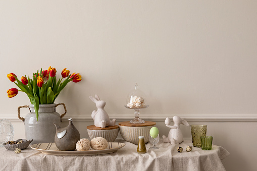 Minimalist composition of living room nterior with copy space, vase with tulips, bread in basket, colorful easter eggs, easter bunny sculpture and personal accessories. Home decor. Template.