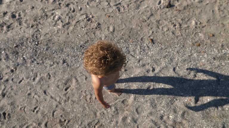 Top Down View of a Cute Toddler Walking on the Sand. Tracking Shot Follows a Little Kid Visiting the Beach for the First Time, Enjoying his Summer Vacation with his Family, Exploring Around