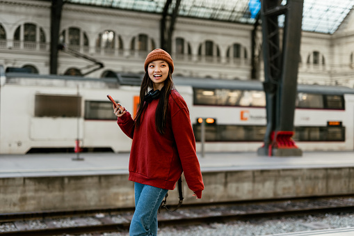 Beautiful young Asian woman standing at modern train station. She is talking with someone using her smart phone. Traveling people concept.