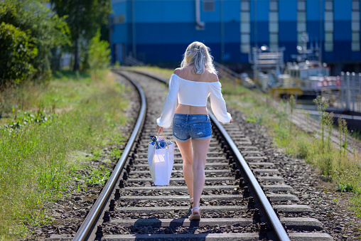 Back view of a blond woman with a white blouse and short jeans and a bag is walking on train tracks
