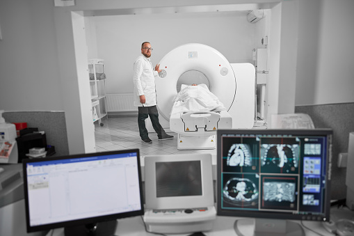Medical computed tomography or MRI scanner. Doctor making MRI, patient lying on couchette. Male specialist wearing white robe and glasses, looking at camera, smiling. Concept of modern diagnostics.