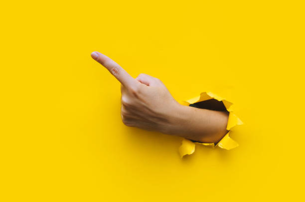 istock The forefinger points to the left up side. Yellow background. Place for advertising. Copy space. The woman's hand came out into the torn paper hole. 1467023163