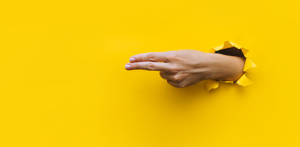 The forefinger and middle fingers point to the left side. Yellow background. Place for advertising. Copy space. The woman's hand was thrust into the torn paper hole.