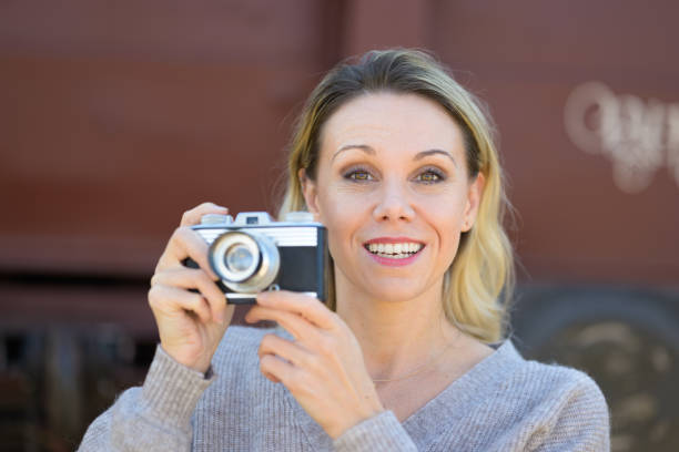 Front view of a smiling middle aged woman holding an old camera wearing a brown sweater in front of an old train wagon