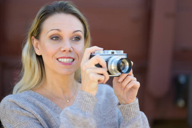 Side view of a smiling middle aged woman holding an old camera wearing a brown sweater in front of an old train wagon