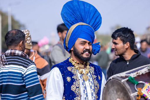 Bikaner, Rajasthan, India - January 2023: Punjabi Bhangra, Portrait of young sikh male in traditional punjabi colorful dress and turban performing bhangra dance with smile in camel festival with selective focus.