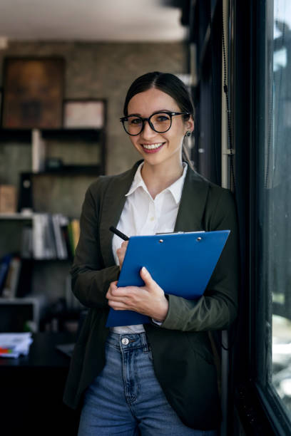 istock Pensive businesswoman in glasses holding file folder looking through window while working in office. Smiling to camera. 1467034372