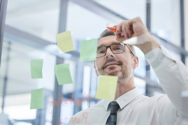 istock Vision, sticky notes and man writing idea in corporate office in strategy meeting, planning and brainstorming. Creative thinking, leader and senior businessman with post it on glass wall in workplace 1467034811