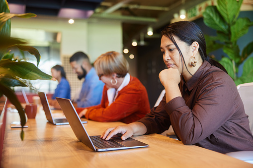 Focused young businesswoman working on a laptop while sitting in a row at a table with a diverse group of colleagues