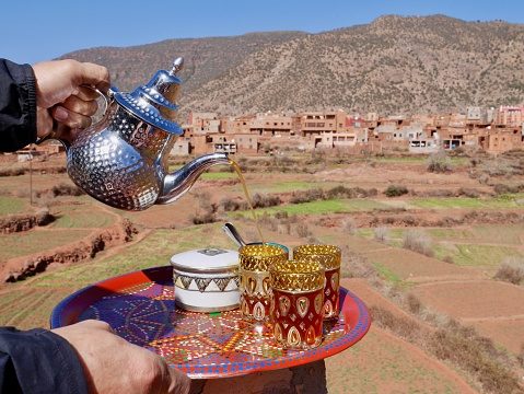 Man pouring mint tea into glasses on colorful tray, Ourika valley and High Atlas Mountains in the background. Morocco.