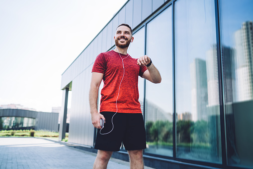 Low angle of cheerful male athlete in sportswear with earphones and smart watch standing on street while preparing for run