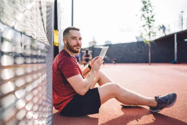 Happy sportsman with short hair and beard sitting near barrier with earphones and browsing tablet while smiling and looking at camera