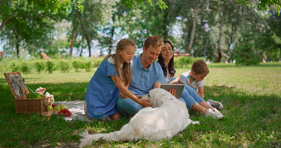 Happy family use tablet on picnic with labrador in summer park. Smiling parents watching laptop sitting with two cheerful kids on blanket on green grass. Tranquil rest with wireless technology outdoor