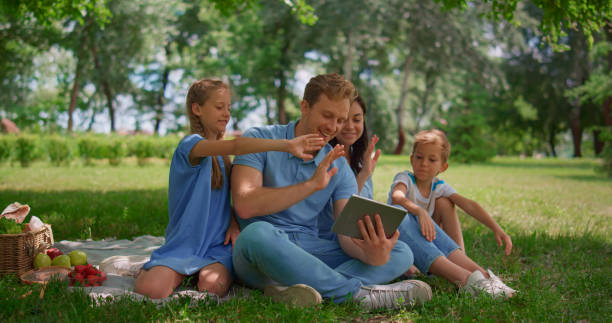 istock Smiling family waving hands communicate using tablet on nature in summer day. 1467045427