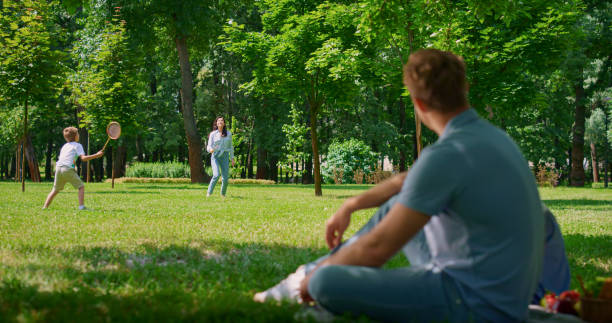 Young father watching badminton game sitting on blanket wearing blue clothes blurred view. Happy son play with mother on green grass summer day. Family activity on weekend in fresh air park.