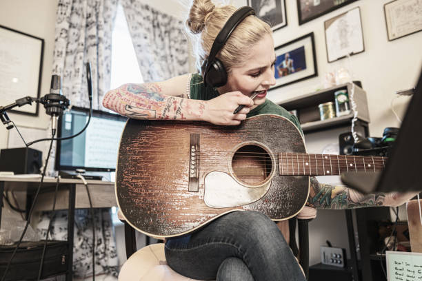 Female Musician practising music on acoustic guitar. She is dressed in casual clothes with sleeve tattoo on both arms.  Interior of home recording studio.