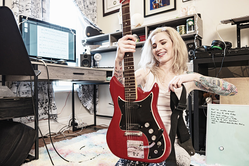 Female Musician practising music on electric guitar. She is dressed in casual clothes with sleeve tattoo on both arms.  Interior of home recording studio.