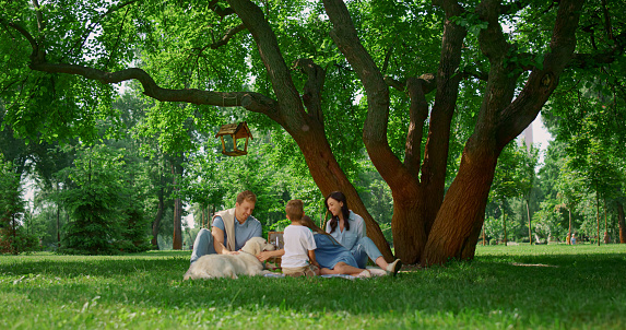 Resting family with dog on picnic under wide tree with bird feeder. Happy people relaxing on park green grass. Parents sitting blanket with two children caressing white labrador on summer meadow.
