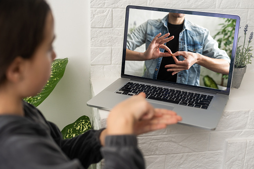 Distant education. Friendly teen girl having online lesson via laptop and showing gesturing in sign language, studying online from home.