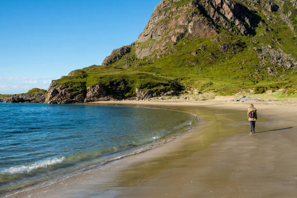 istock Young woman with a backpack walks along an empty sandy ocean beach. Town and mountains. Beautiful nature landscape. Lonely girl. Wanderlust. Travel, adventure, lifestyle. Explore North, Norway 1467062737
